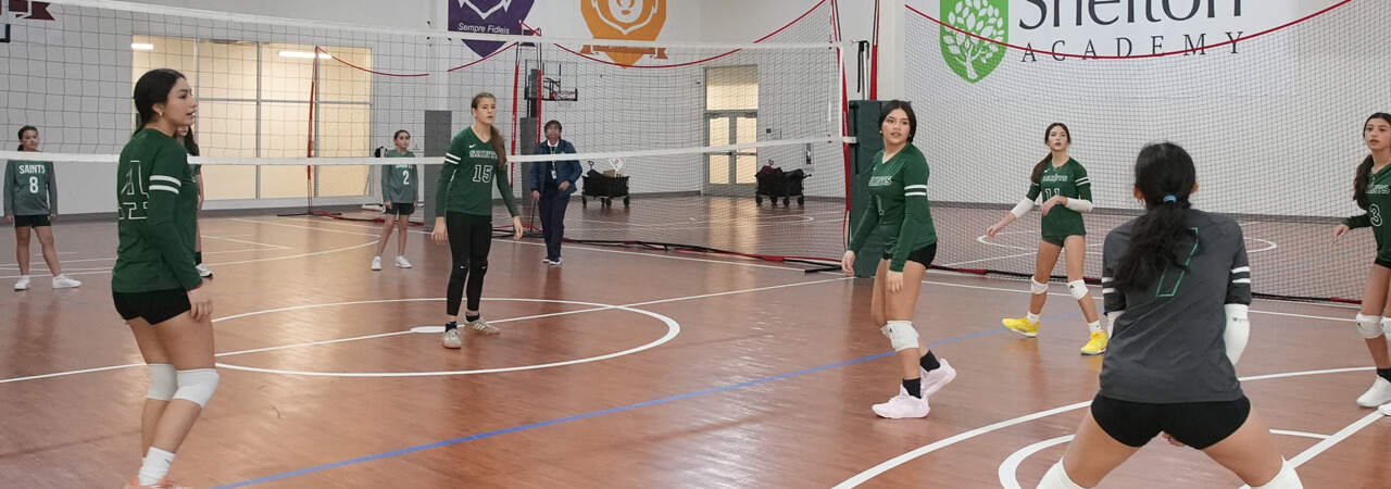Female volleyball players in green jerseys preparing for a serve on a polished indoor court.