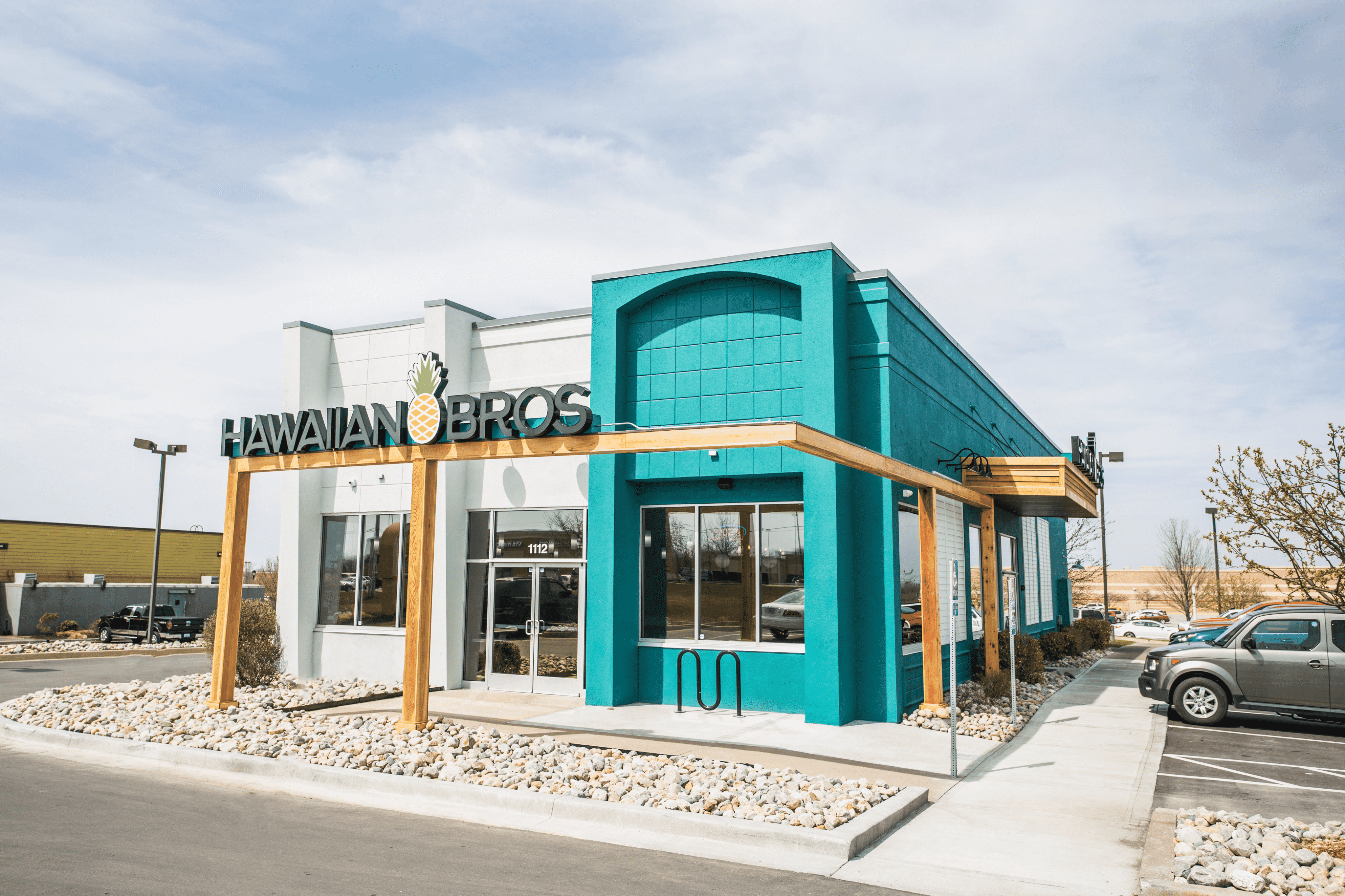 Outside of a Hawaiian Bros location with a large sign and teal walls. 