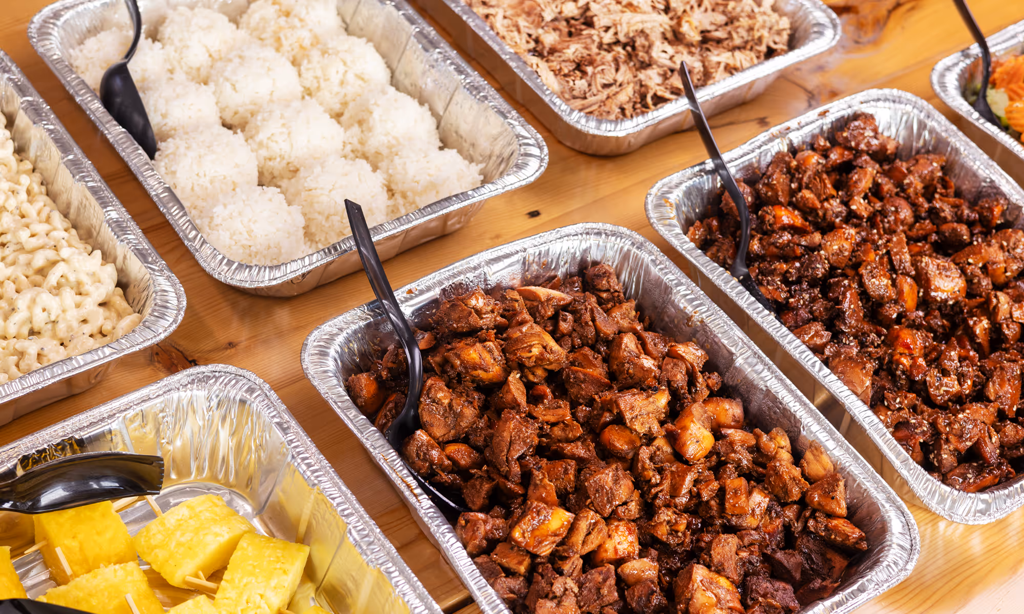 Trays of Hawaiian Bros catering with various protein options, pineapple, and white rice.