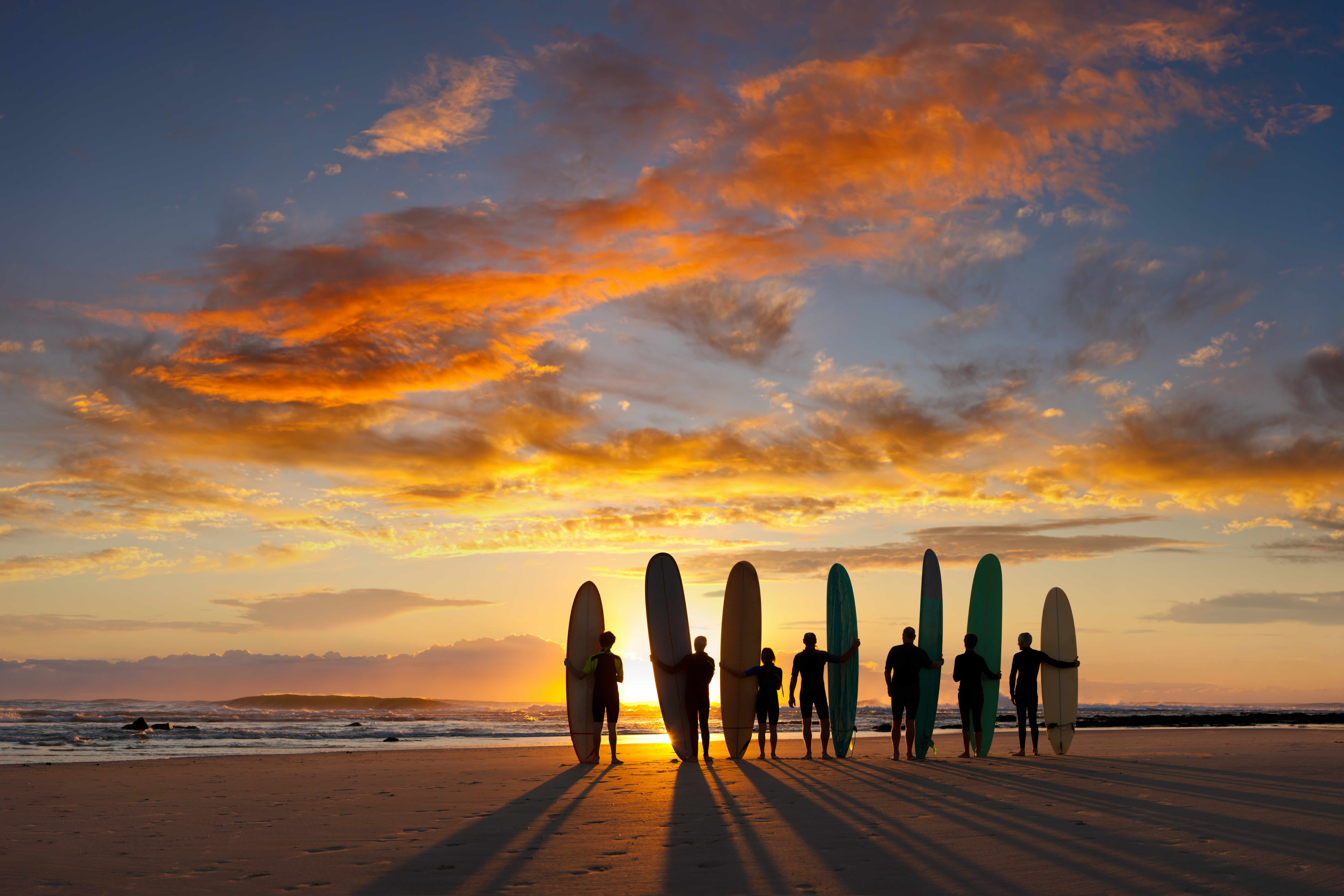The silhouettes of a group of surfers standing on the beach with their long boards at sunset.