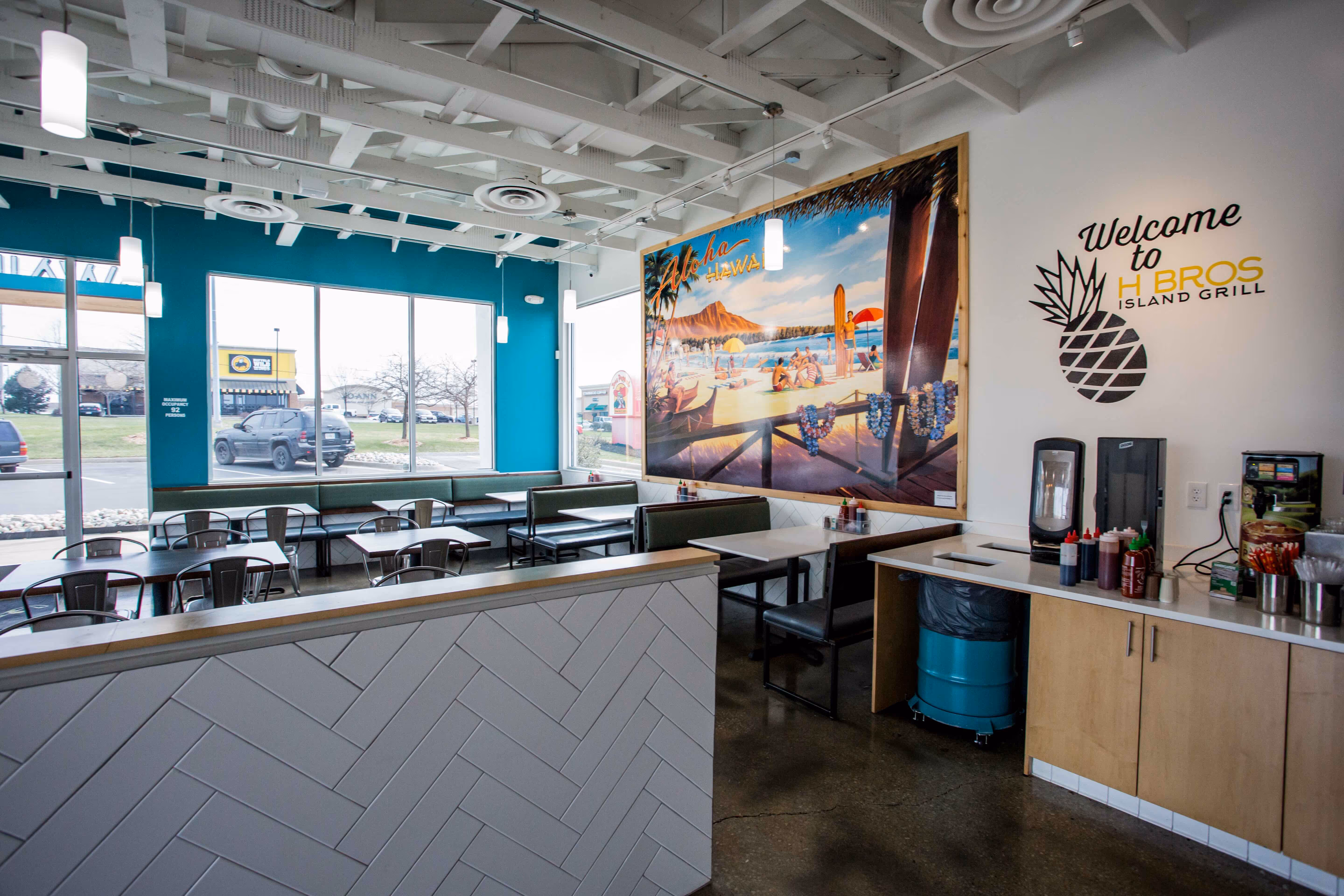 Inside of a Hawaiian Bros location with a seating area, a large framed picture of Hawaii, and the utensil and condiment station.