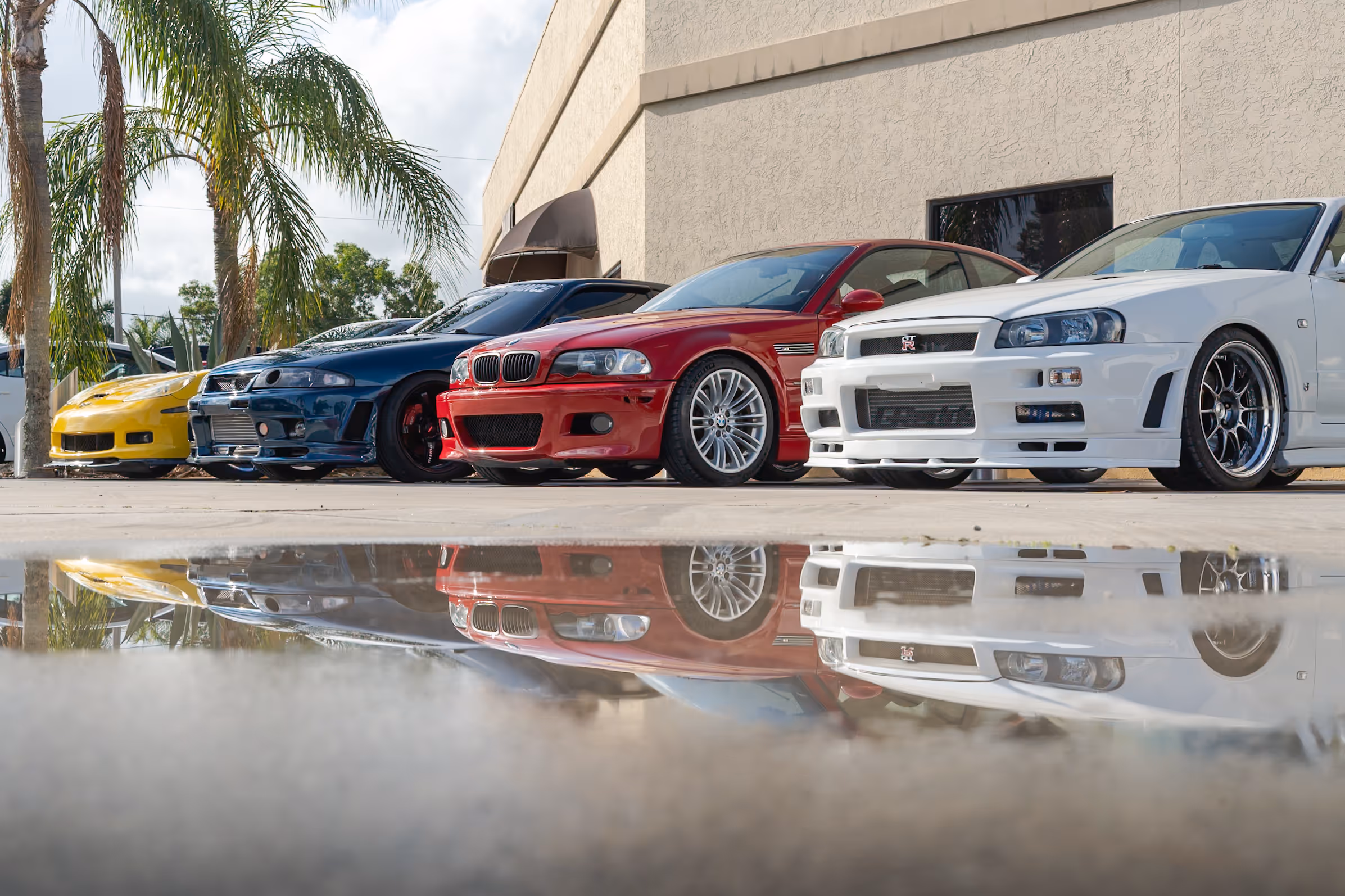 Four colorful sports cars parked with reflection on wet ground near palm trees
