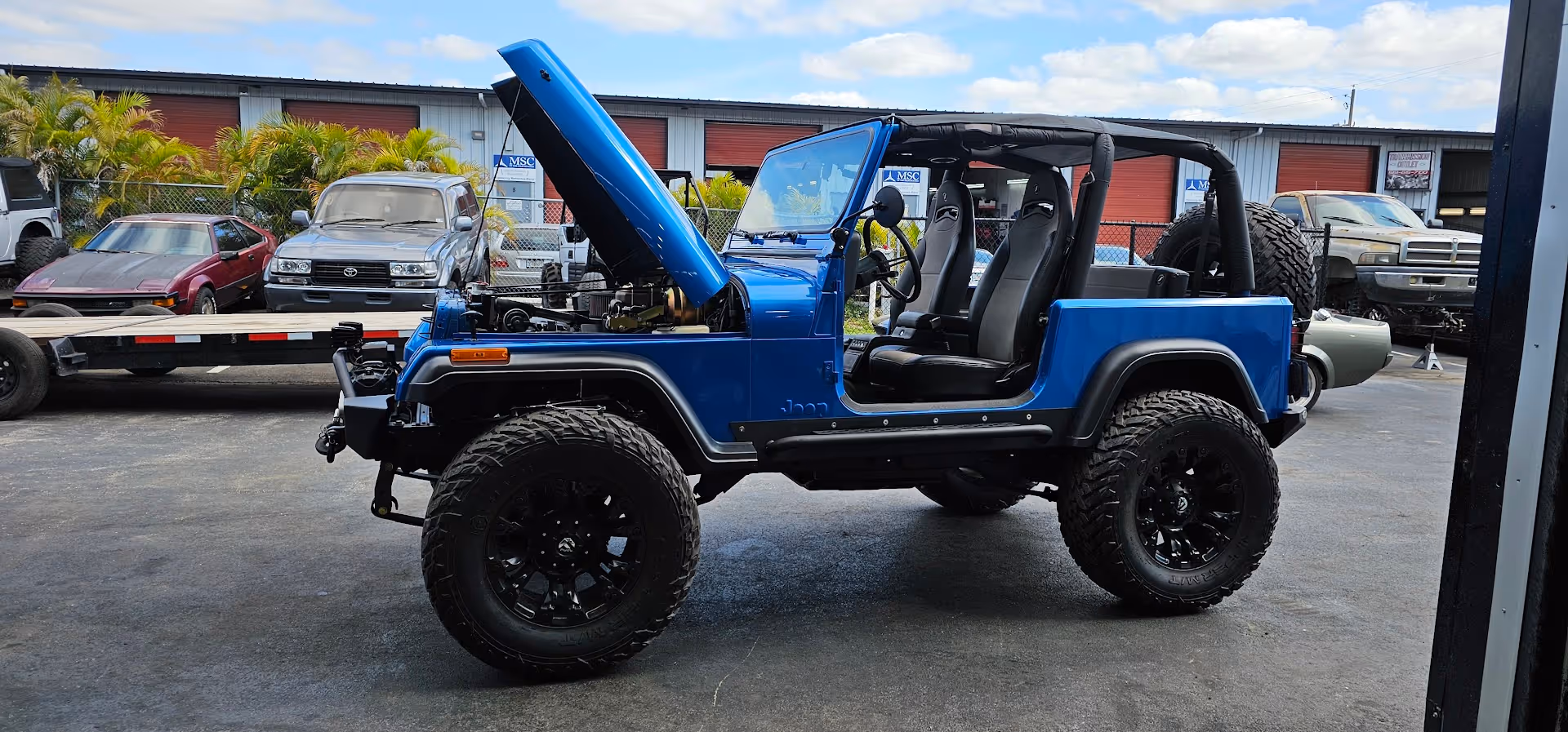Blue Jeep with open doors and hood, large off-road tires in workshop