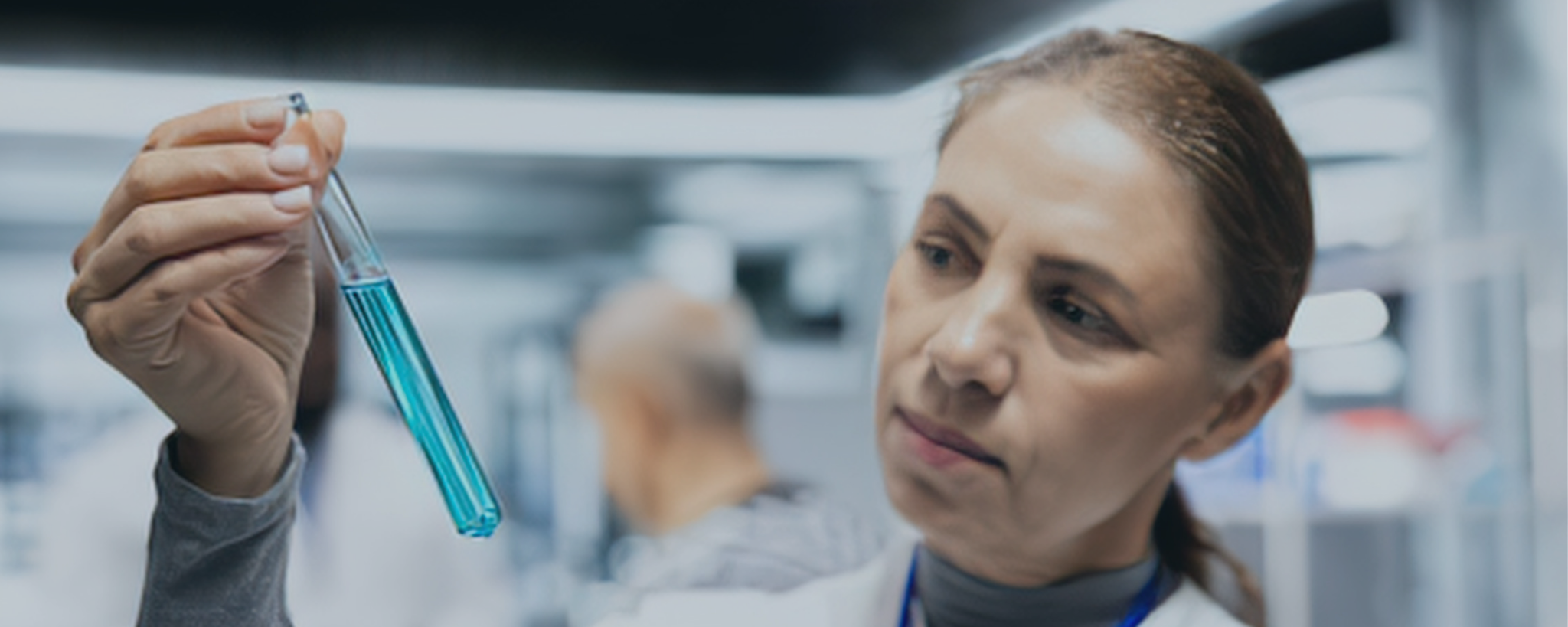 Eine Frau im Laborkittel betrachtet ein Reagenzglas mit türkisfarbener Flüssigkeit in einem Labor.