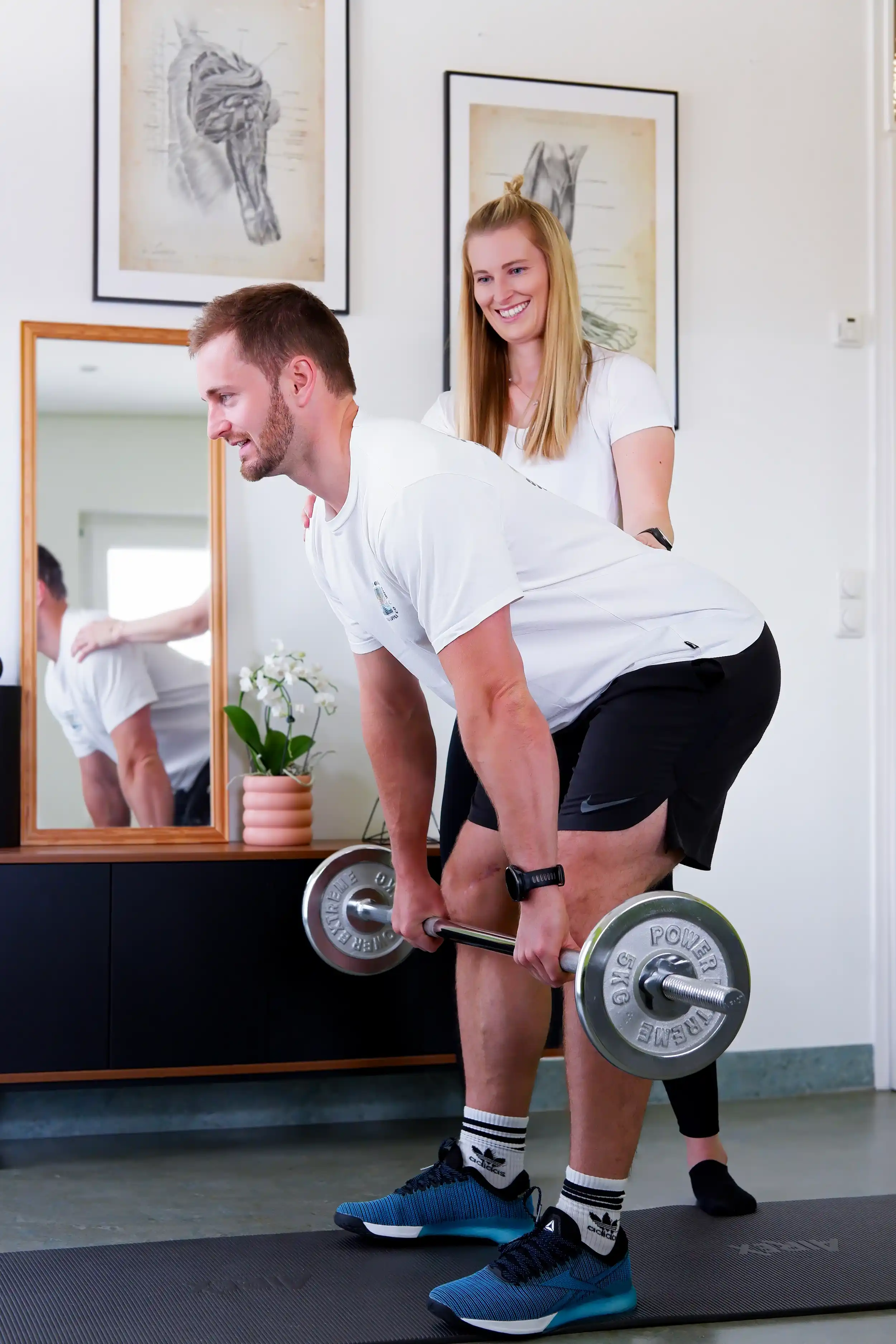 Sportphysiotherapeutin begleitet einen Patienten bei einer Kraftübung mit Langhantel in der Praxis