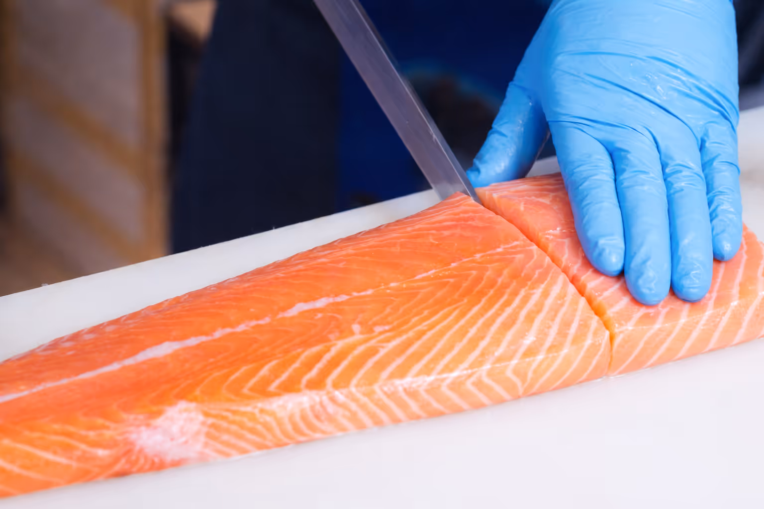 Hand wearing blue glove slicing a fresh salmon fillet with a knife on a white cutting board.