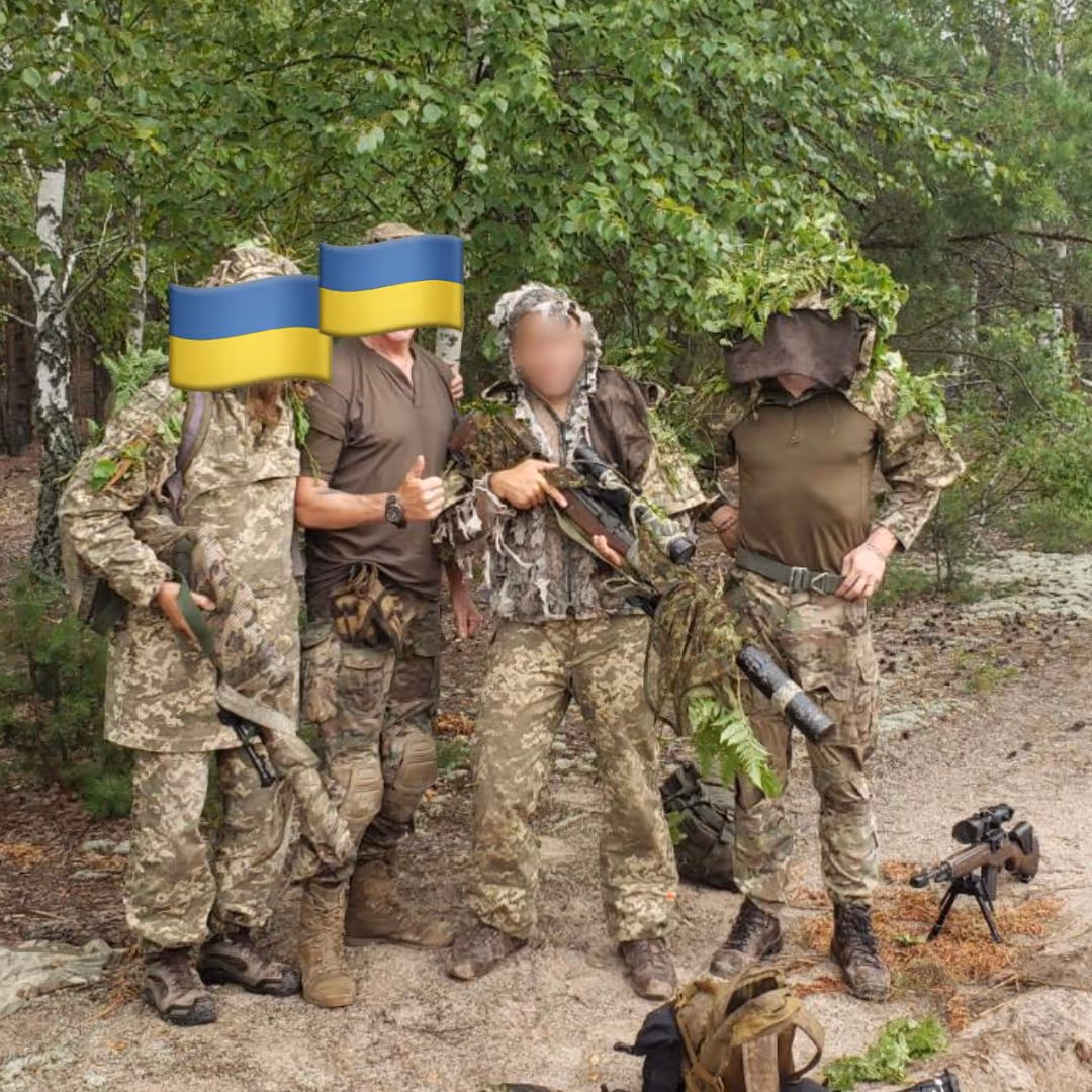 Four soldiers in camouflage uniforms posing outdoors among trees, with two faces covered by Ukrainian flag emojis and two faces blurred; one soldier holds a camouflaged rifle, and another has branches on his helmet.