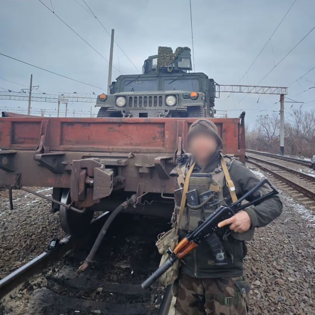 Soldier in camouflage and tactical gear holding a rifle standing in front of a military vehicle on a railway flatcar.