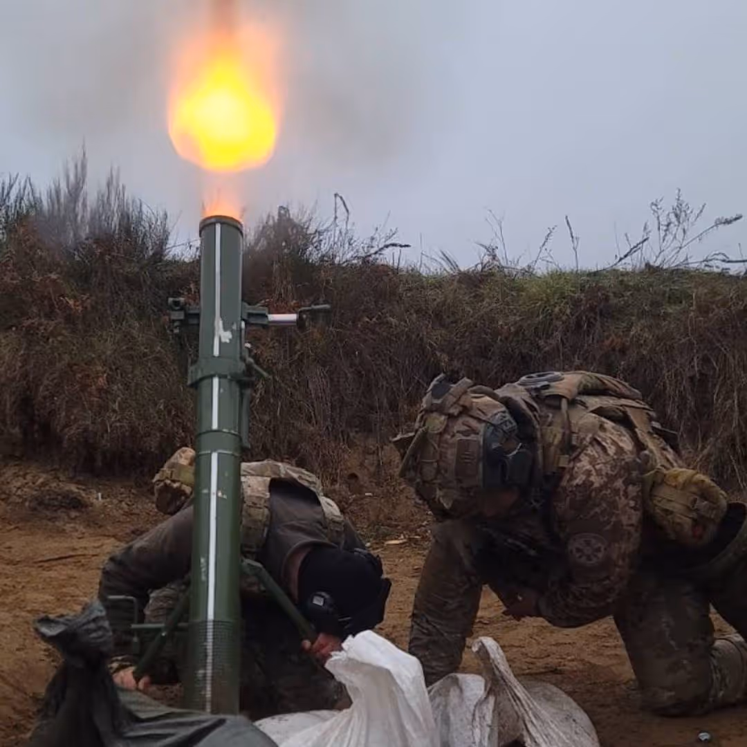 Two soldiers in camouflage uniforms operating a mortar launching a bright fiery projectile outdoors.