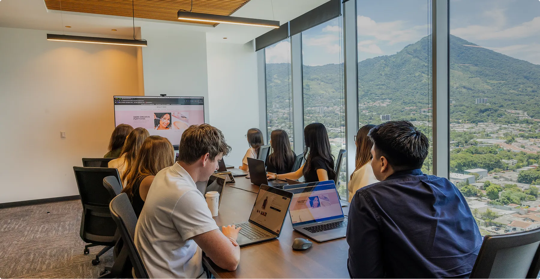 Team of professionals seated around a conference table in a modern office with a mountain view, working on laptops and viewing a screen presentation.