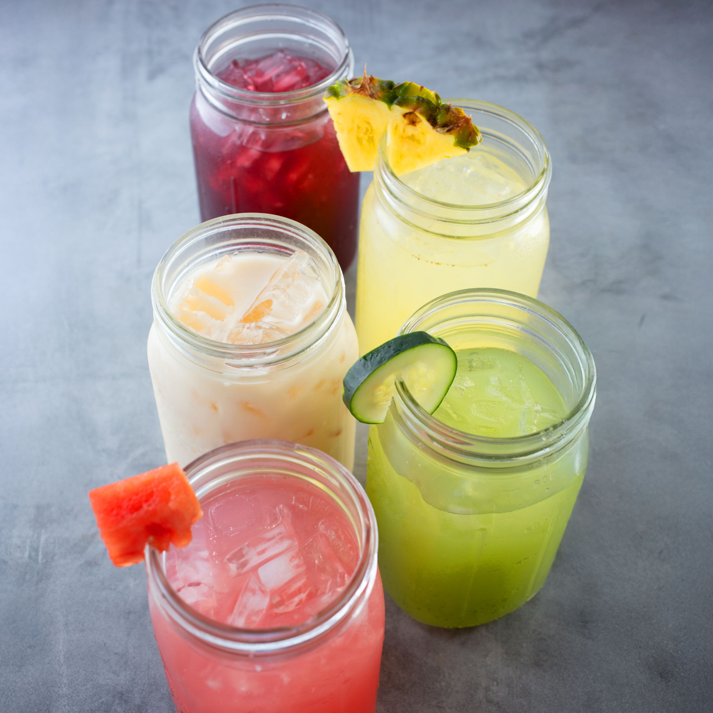 Five colorful drinks in mason jars on a gray surface. Each jar is uniquely garnished with fruit, including pineapple, cucumber, and watermelon.
