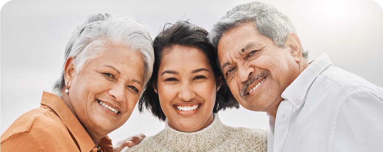 Three people, two older adults and a younger woman, stand close together outdoors, smiling warmly at the camera with a bright, light background.