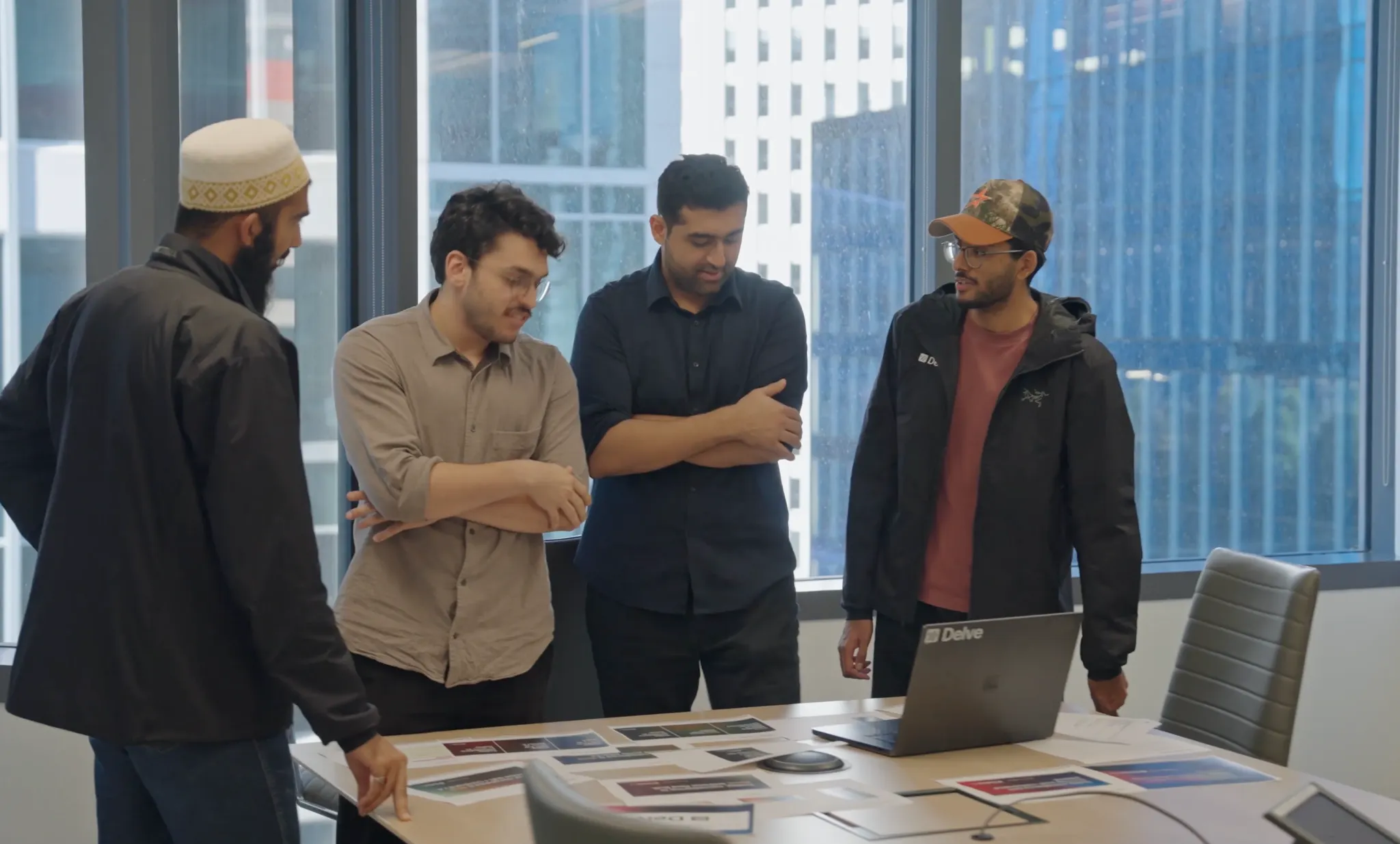 Four men standing around a table with printed materials and a laptop in a modern office with large windows.