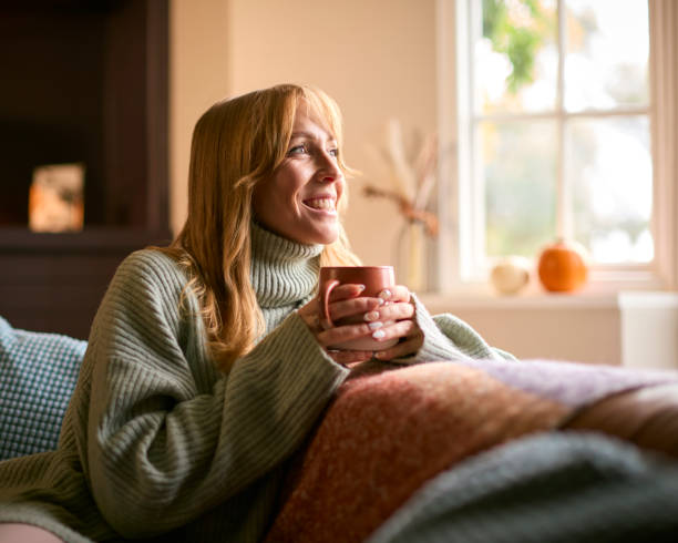 Smiling Woman At Home Wearing Winter Jumper With Warming Hot Drink Of Tea Or Coffee In Cup Or Mug Smiling Woman At Home Wearing Winter Jumper With Warming Hot Drink Of Tea Or Coffee In Cup Or Mug woman holding coffee during fall stock pictures, royalty-free photos & images