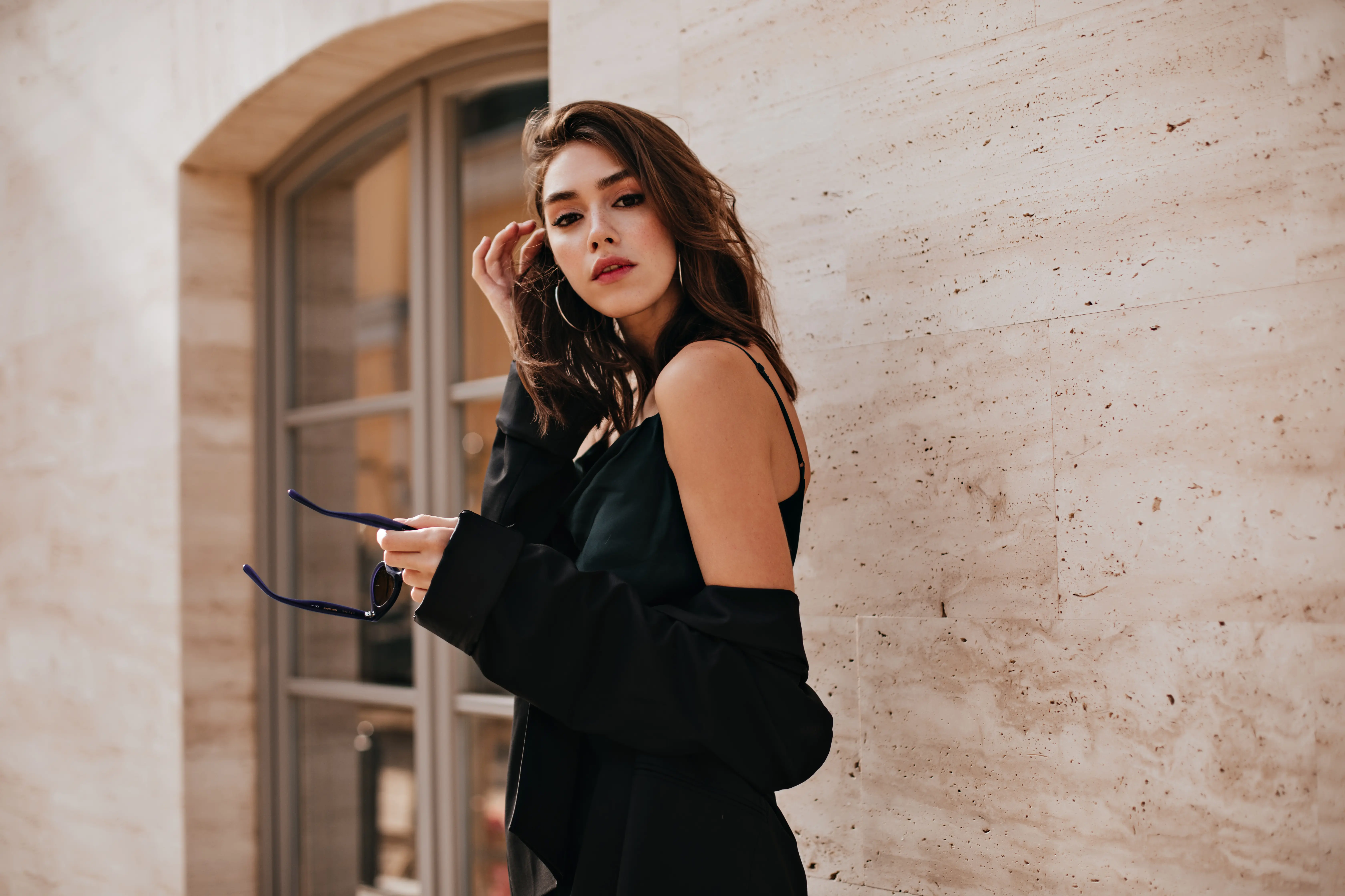Young woman with loose brown hair wearing hoop earrings and a black off-shoulder outfit, holding sunglasses and standing against a beige stone wall near a window.