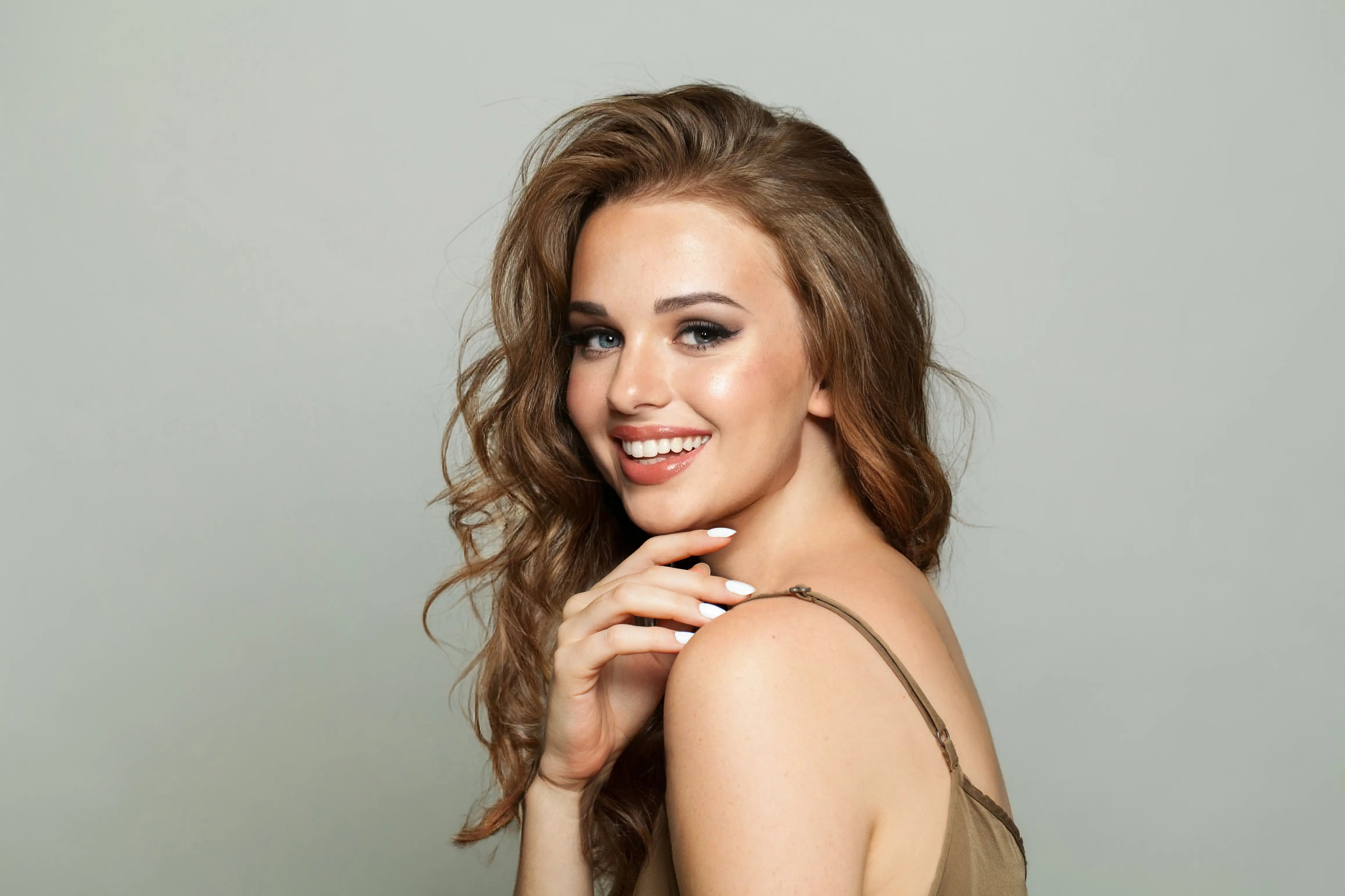 Smiling woman with long wavy hair and natural makeup posing with hand near her chin against a plain background.