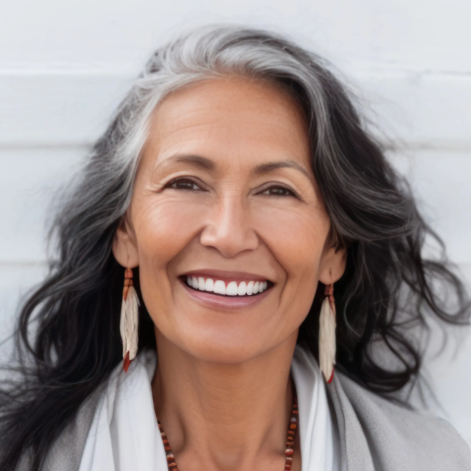 Smiling middle-aged woman with long black and gray hair, wearing feather earrings and a beaded necklace.