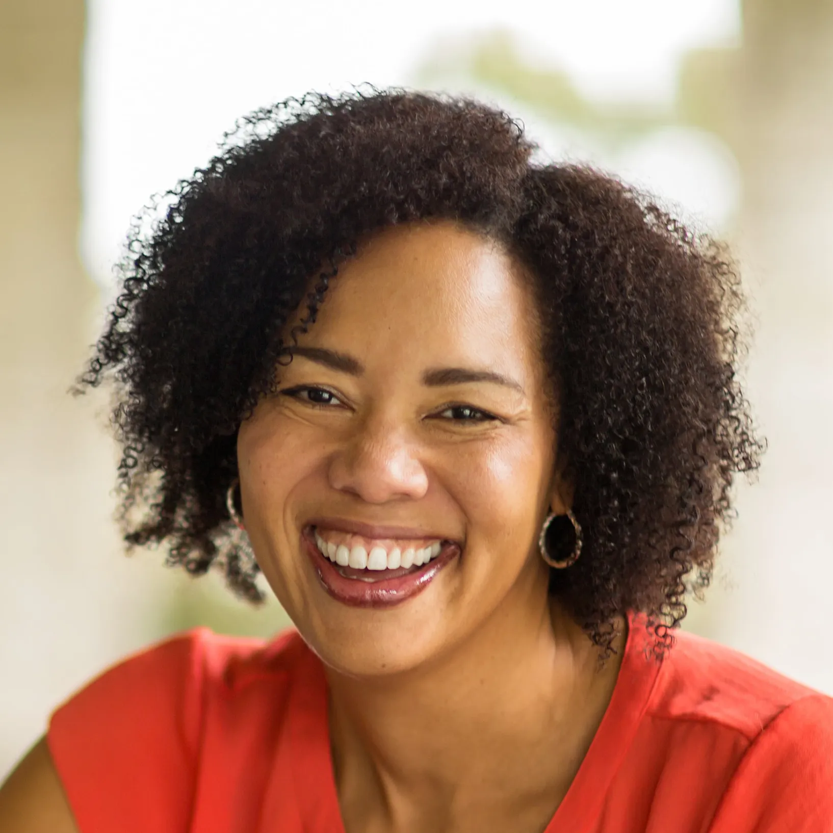 Smiling woman with curly black hair wearing a red top and hoop earrings.