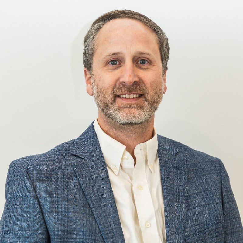 Smiling man with light brown hair and beard wearing a dark suit jacket and white shirt standing in front of a wooden panel background.