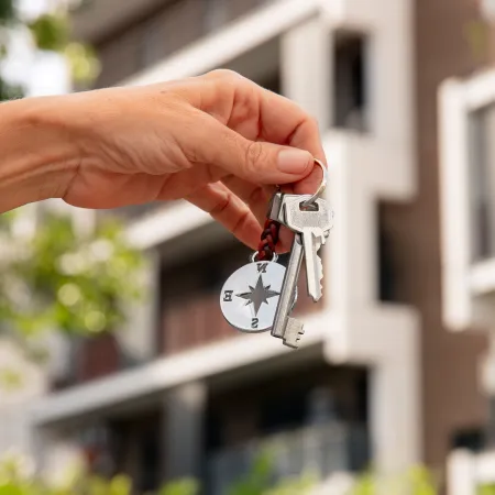 Hand holding a set of keys with a compass keychain in front of a blurred building.