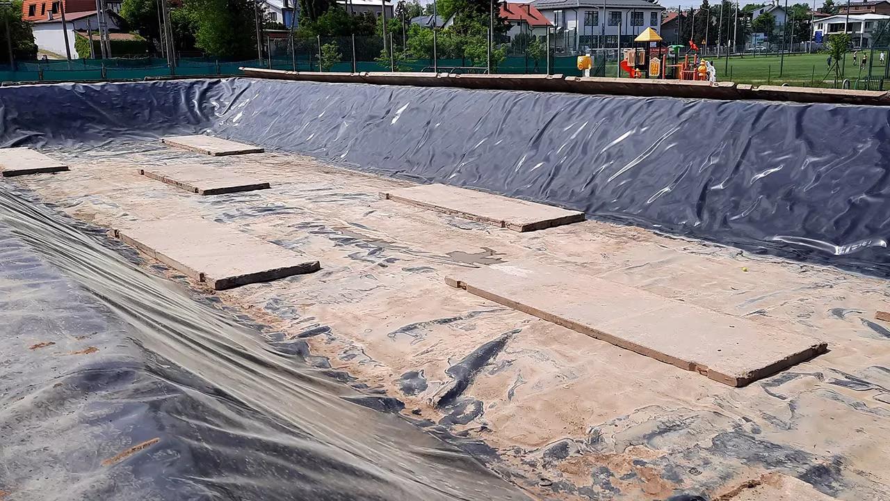 Pusty basen wyłożony czarną folią, z płytami betonowymi na dnie, otoczony przez domy i plac zabaw w tle.