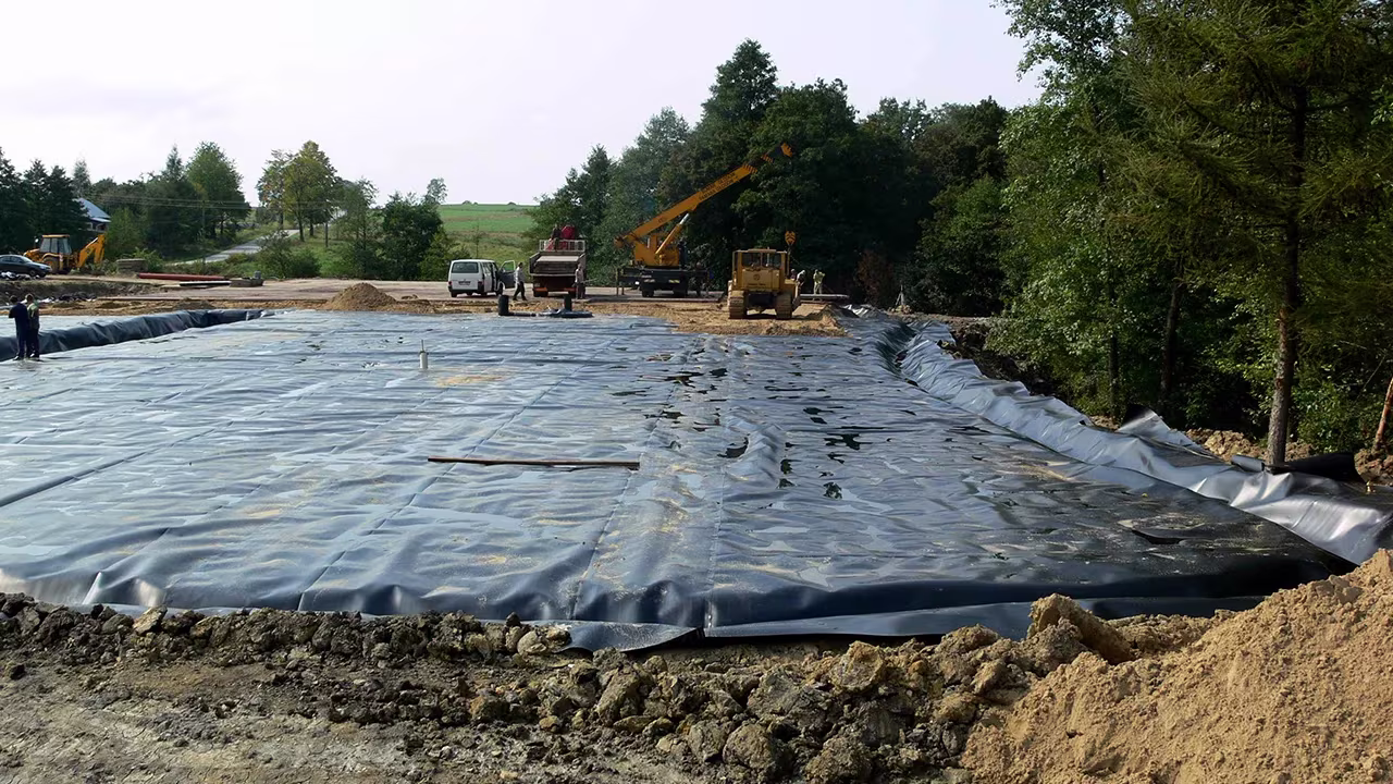 Plac budowy pokryty czarną folią do izolacji ziemi, z maszynami budowlanymi i pracownikami w tle, otoczony drzewami.