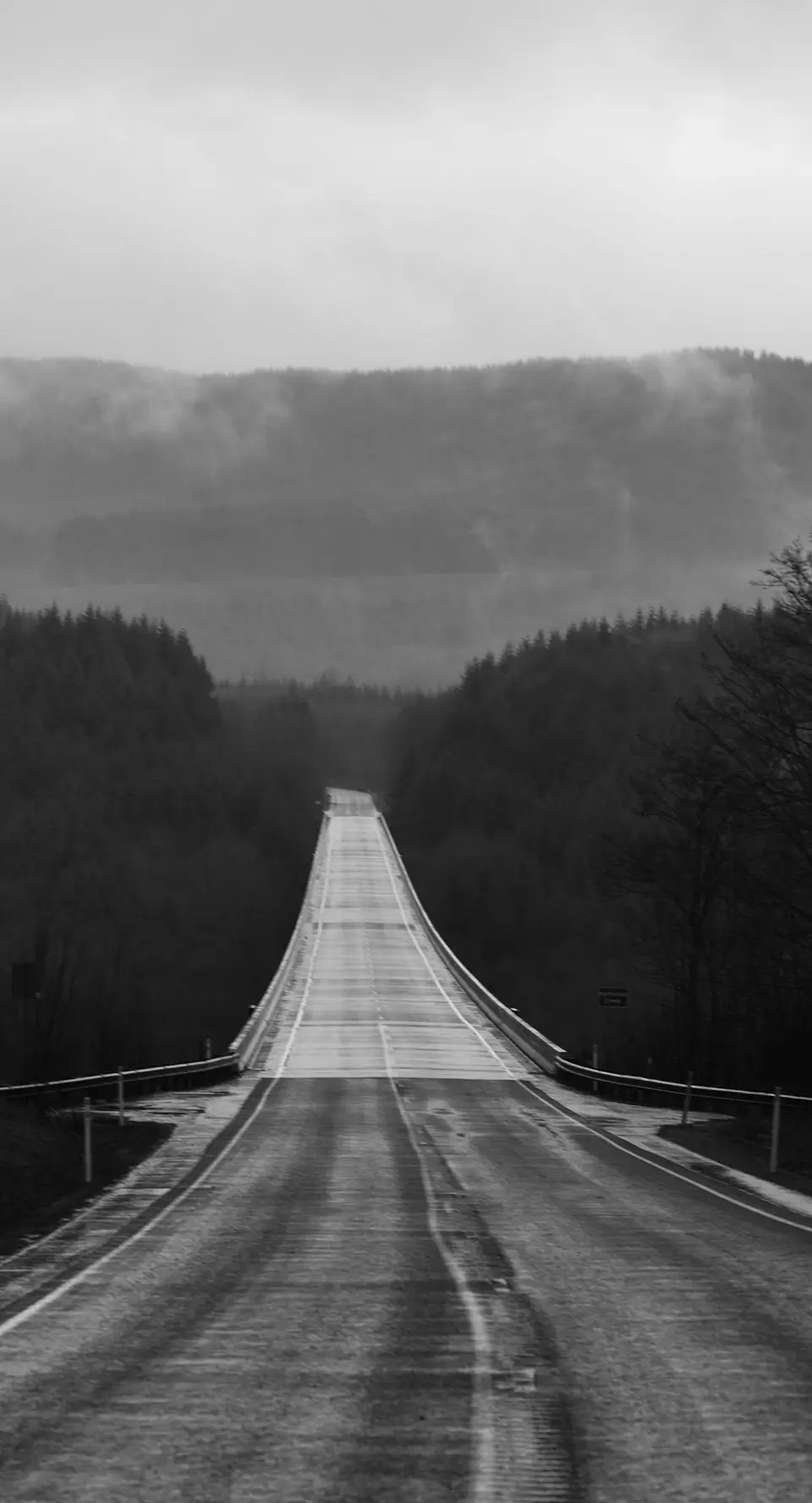 Empty two-lane road stretching into foggy, forested hills under a cloudy sky.