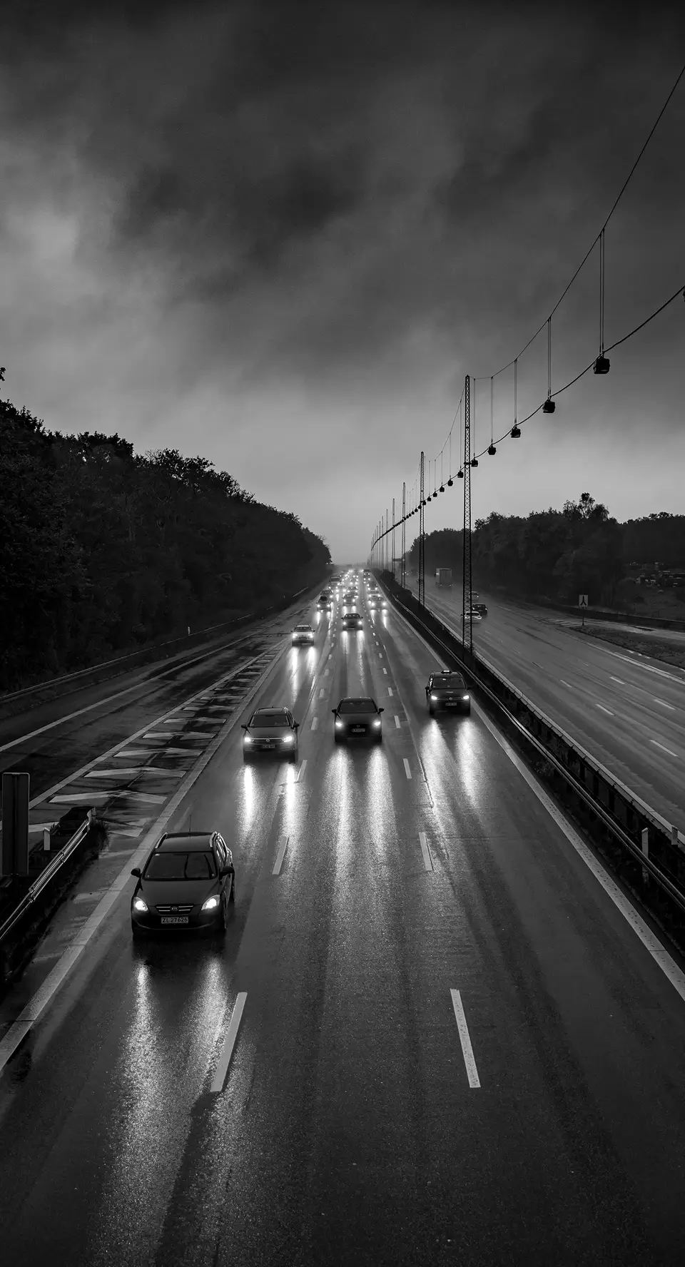 Cars with headlights on driving on a wet multi-lane highway under a cloudy sky at dusk.