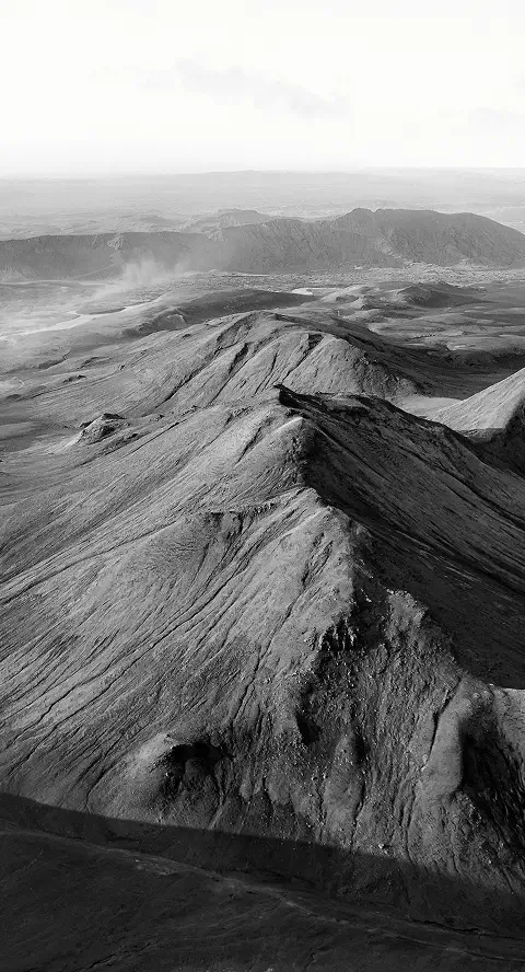 Black and white aerial view of rugged volcanic terrain with peaks and ash-covered slopes.