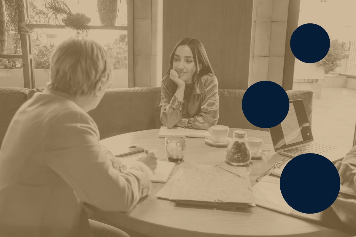 A woman facing the camera sitting at a table in a cafe for a job interview with another person who is facing away from the camera