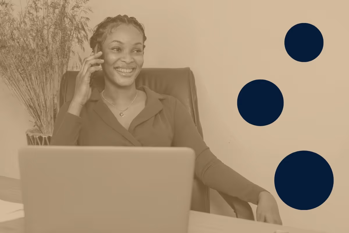 A smiling woman sitting at a desk and holding a smartphone up to her ear