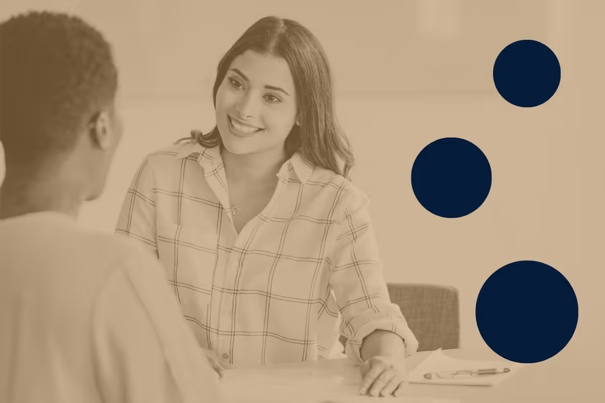 A woman facing the camera and smiling while listening to another woman, who is facing away from the camera, speak