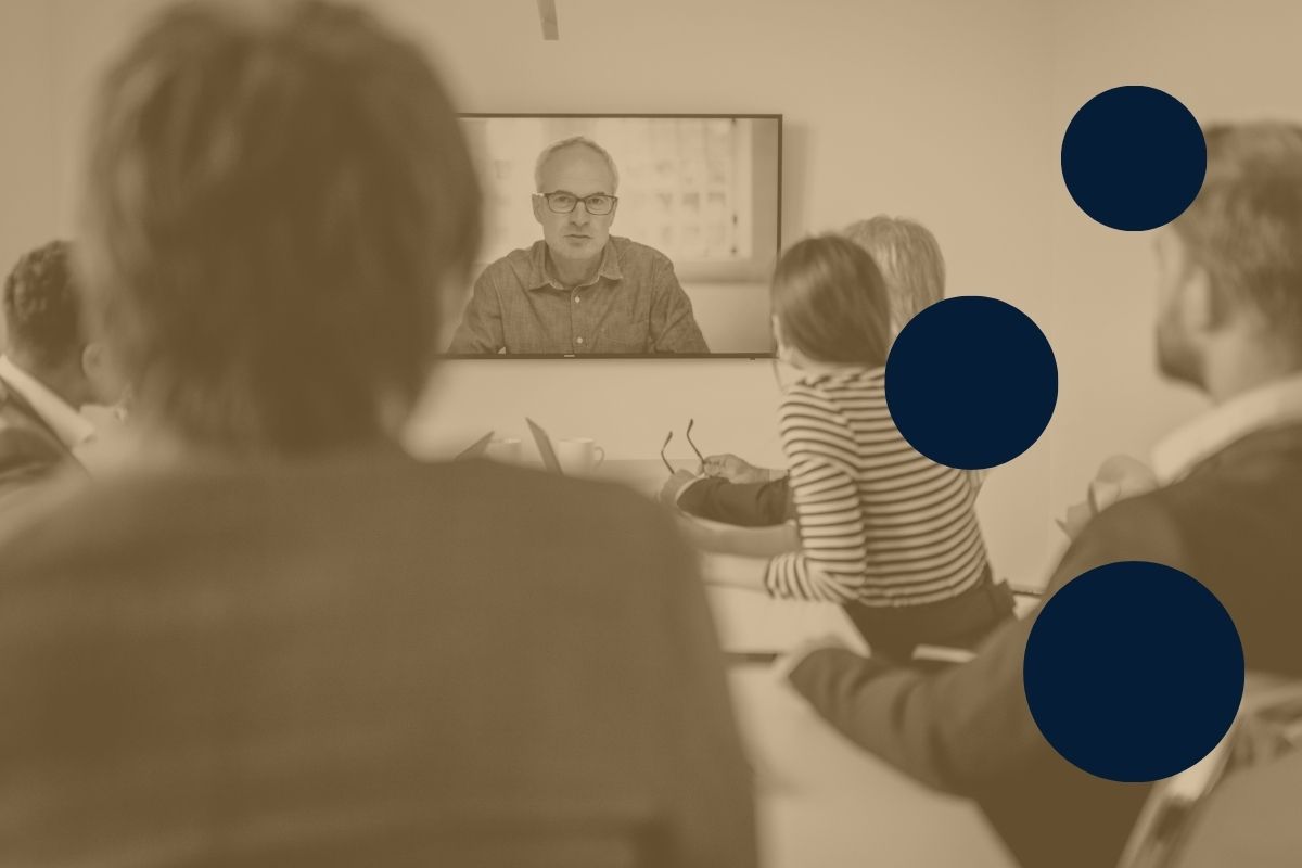 A group of professionally-dressed people sitting around a conference table and looking at a large, wall-mounted screen displaying a man on a conference call who is speaking to the group