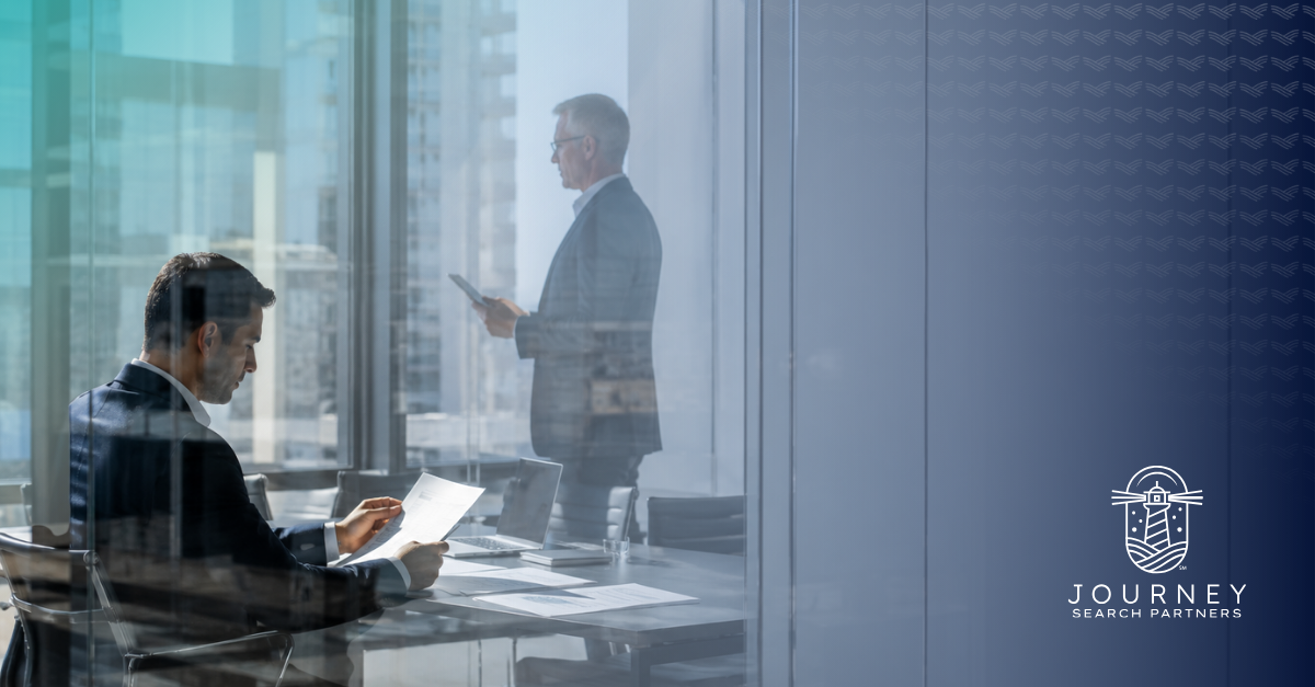 Two businessmen in a glass-walled office, one reviewing documents at a table and one standing by the window, representing what sets good managers and great leaders apart.