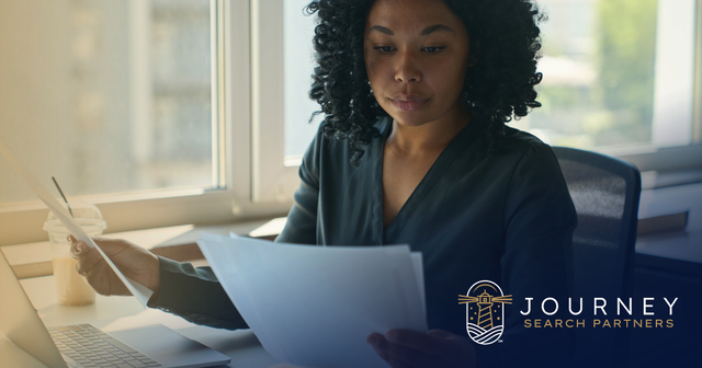 A professional woman at a sunny window desk reviews printed documents beside her laptop. Journey Search Partners logo.