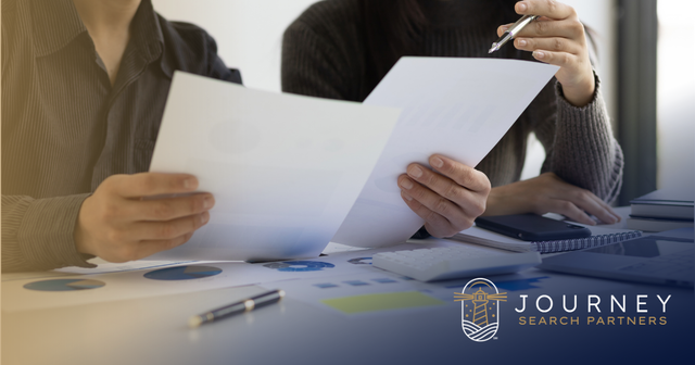 Two colleagues review printed documents over a desk covered in charts and reports. Journey Search Partners logo.