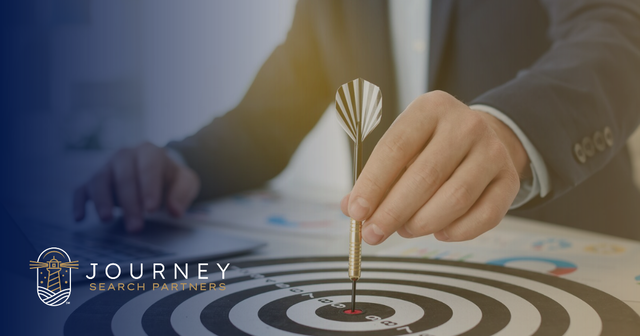 A businessman places a dart in the bullseye of a dartboard on his desk. Journey Search Partners logo.