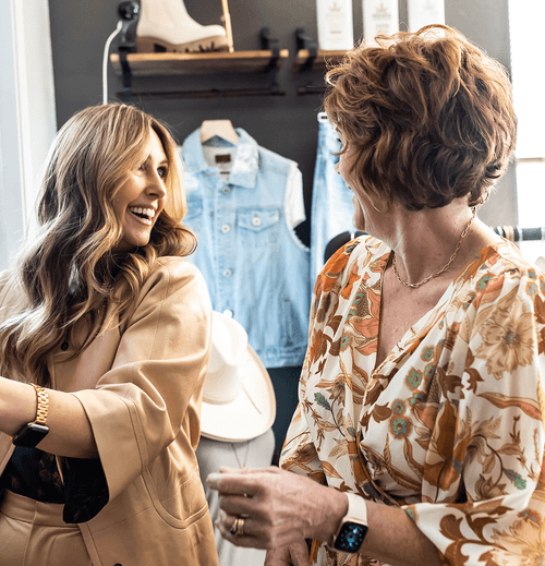 Two smiling women talking inside a clothing store with denim jackets and hats on display behind them.