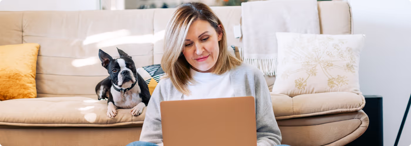 Women sitting on the floor looking at her laptop with a dog sitting on the couch directly behind her