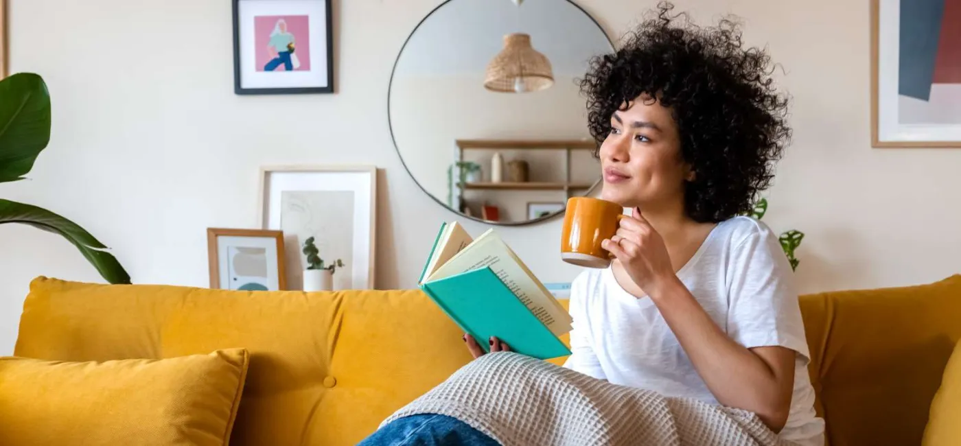 Young woman sitting on the couch drinking out of mug and reading a book