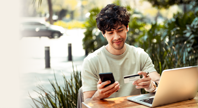 Younger male sitting at a table with a laptop looking at his credit card and mobile phone