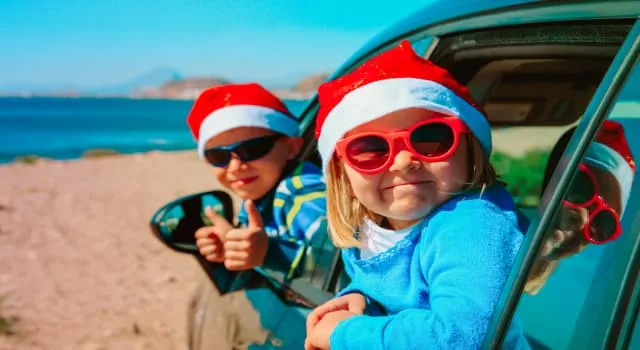 Two young children smiling with the thumbs up in Christmas hats by the beach