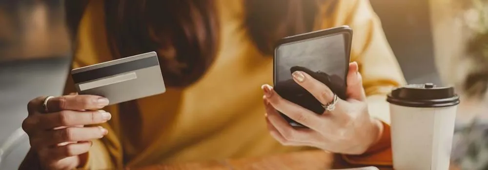 A woman is holding a credit card in one hand and a smartphone in the other, appearing engaged in a financial transaction.