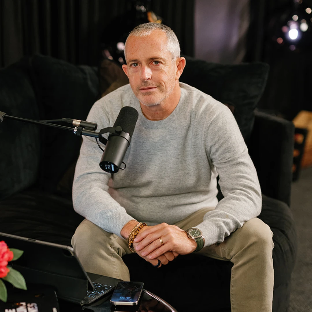 Two people wearing headphones sitting and talking into microphones in a modern podcast studio with a large illuminated G on the wall.