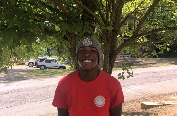 young black male teen smiling in a red shirt and grey beanie in front of a tree