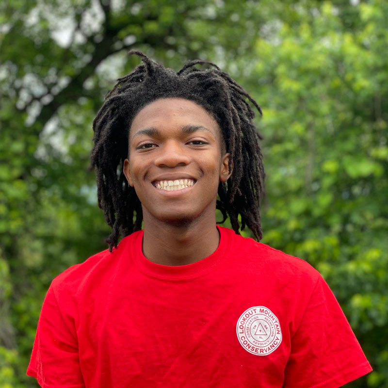 Headshot of a black teenage man with dreds in front of a forested background.