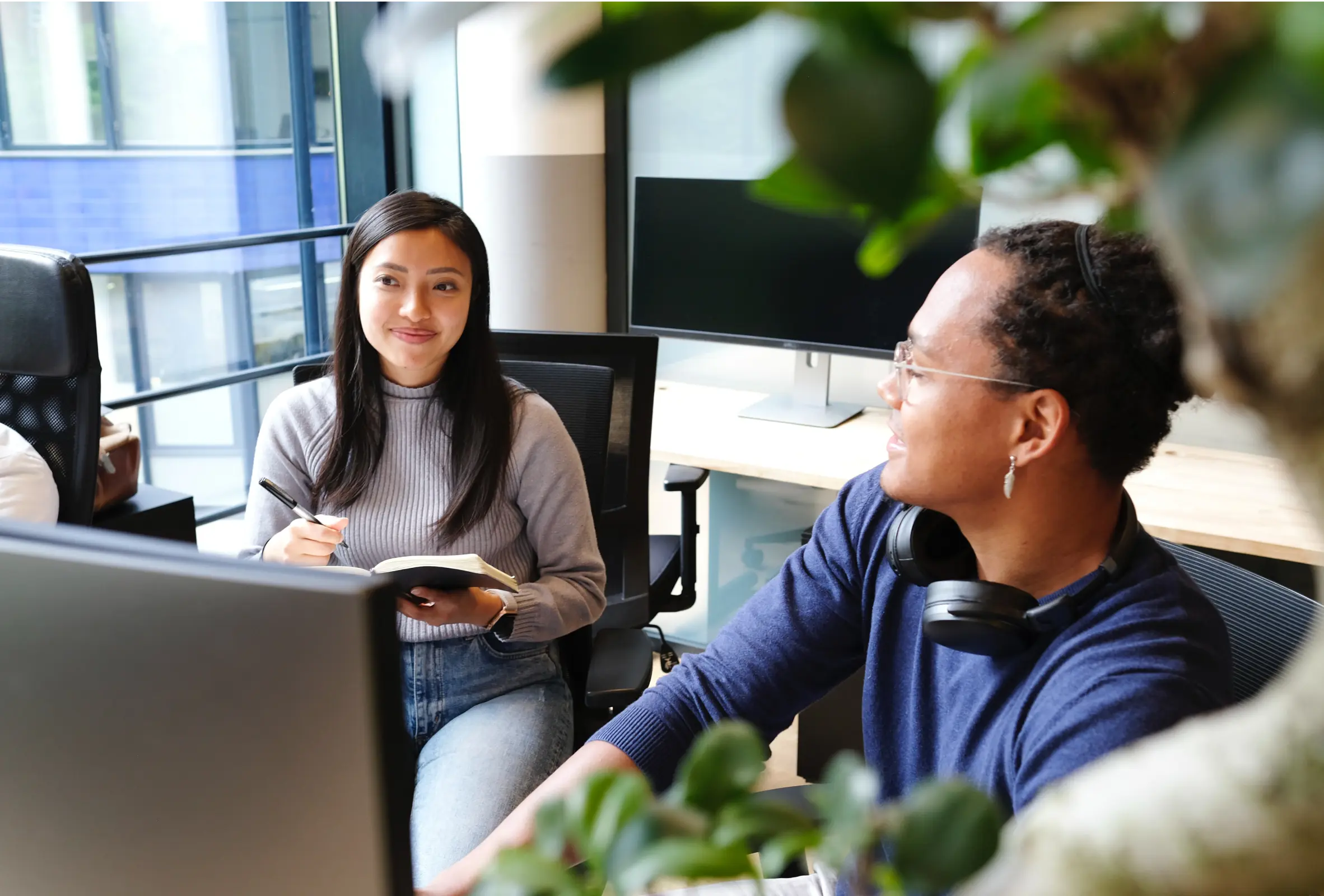 A focused woman in a grey sweater and blue jeans sits at a desk, looking to her left while holding a pen and notebook. Beside her, another woman with glasses and headphones around her neck, also seated at a desk, looks to her right. Both are in an office setting with computer monitors visible in the background and foreground, and plants adding a touch of greenery.