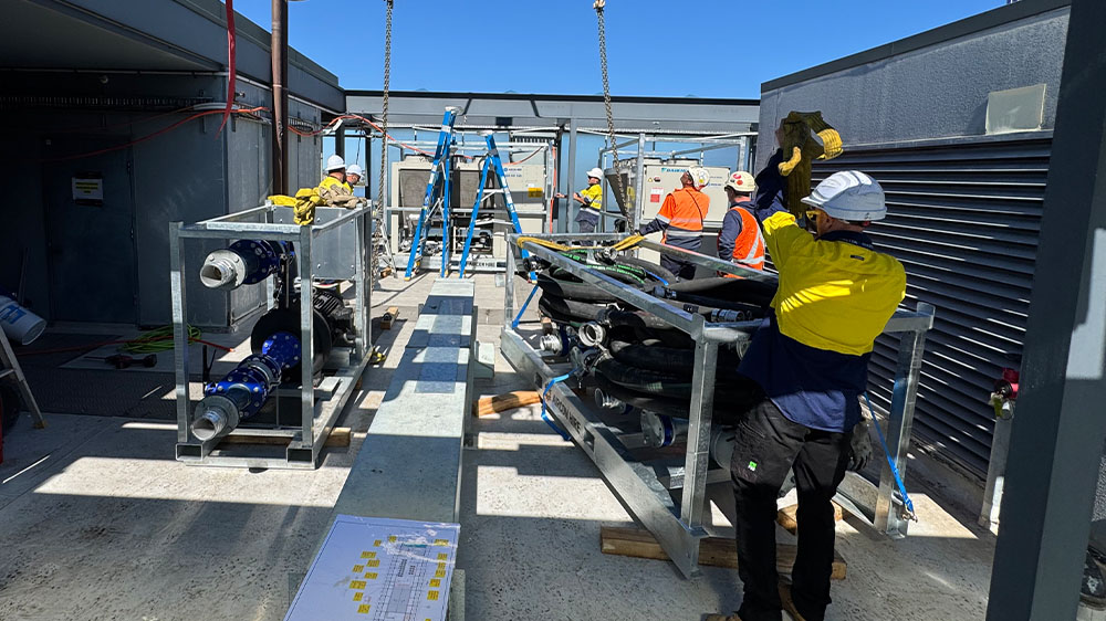Workers wearing safety helmets and high-visibility clothing assembling industrial equipment on a rooftop under a clear blue sky.