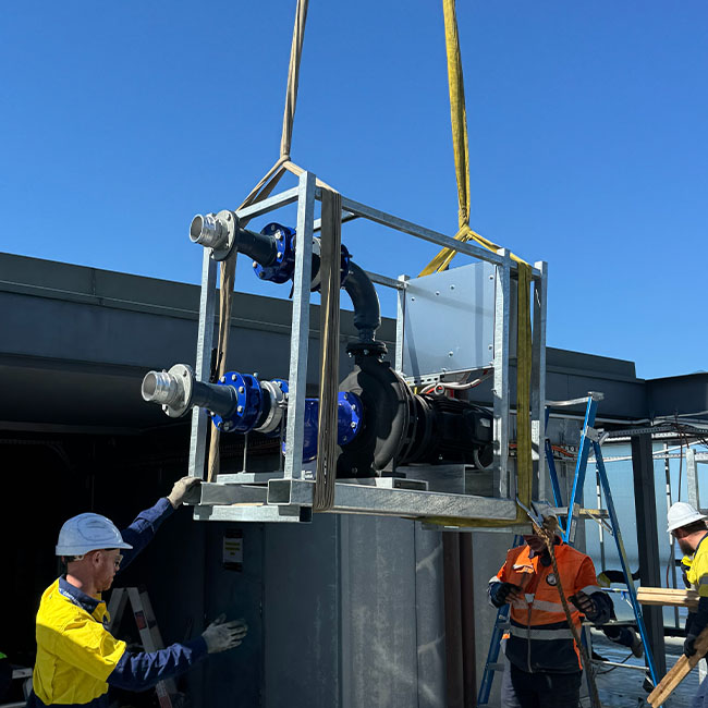 Three workers in safety gear guiding a large metal-framed pump being lifted by straps against a clear blue sky.