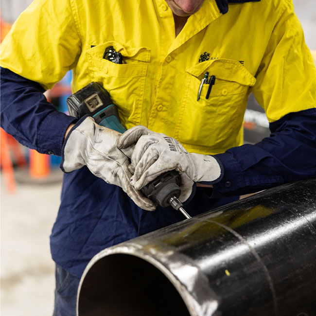Worker wearing yellow and blue shirt with gloves using a power drill on a large black metal pipe.